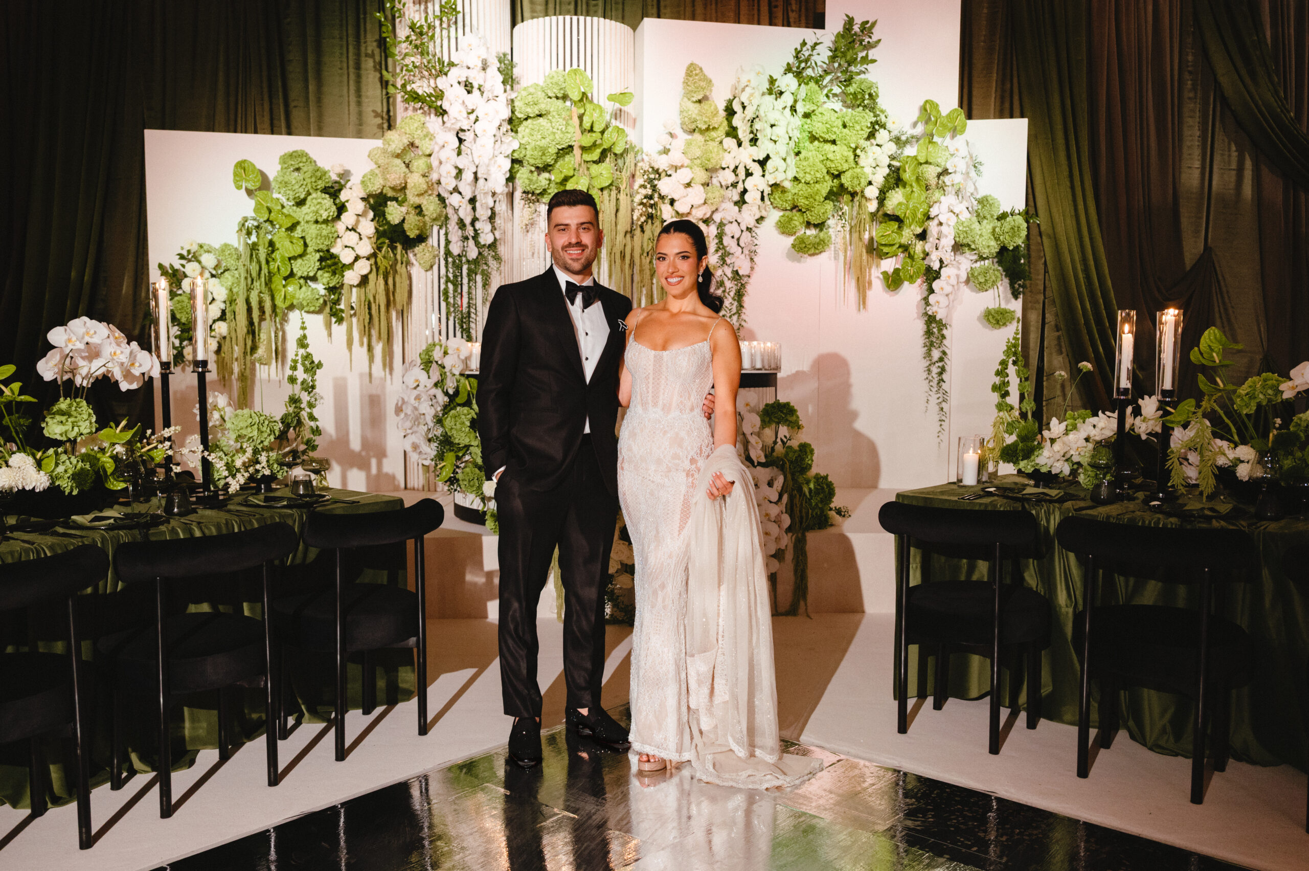 Valerie and Cosimo stand together on a glossy aisle amid white flowers, greenery, candles, and black reception tables.
