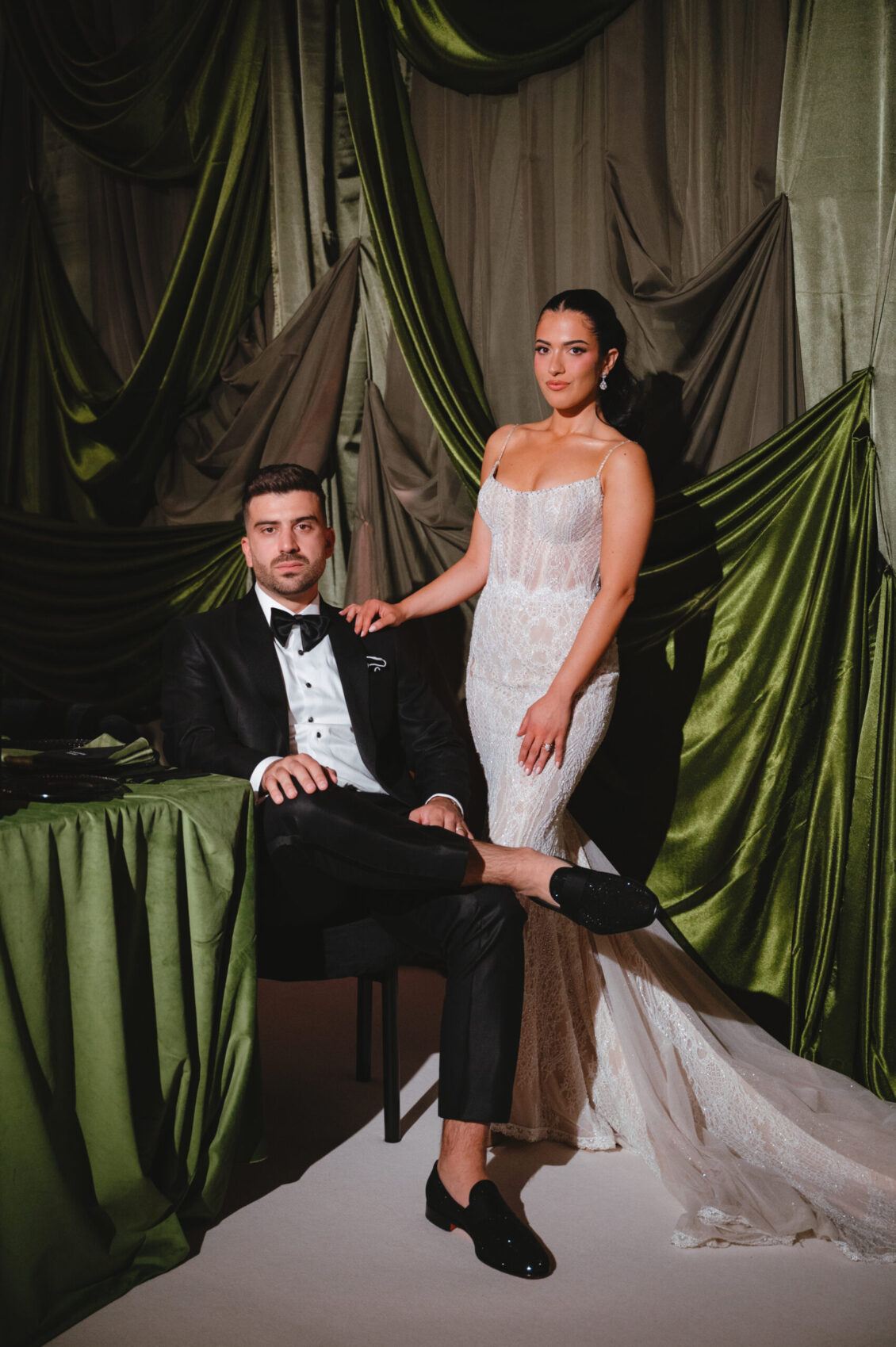 Cosimo sits beside an olive-green table while Valerie stands in her fitted gown with her sheer train spread across the floor.