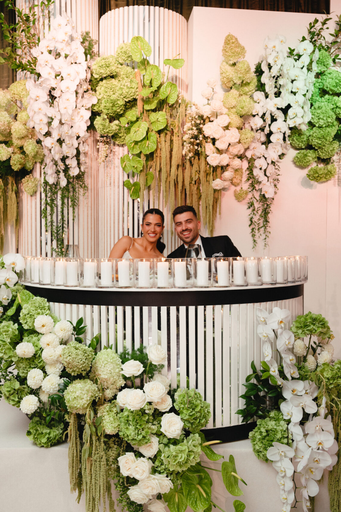 Valerie and Cosimo smile behind a curved black-and-white sweetheart table lined with candles and surrounded by hanging flowers.