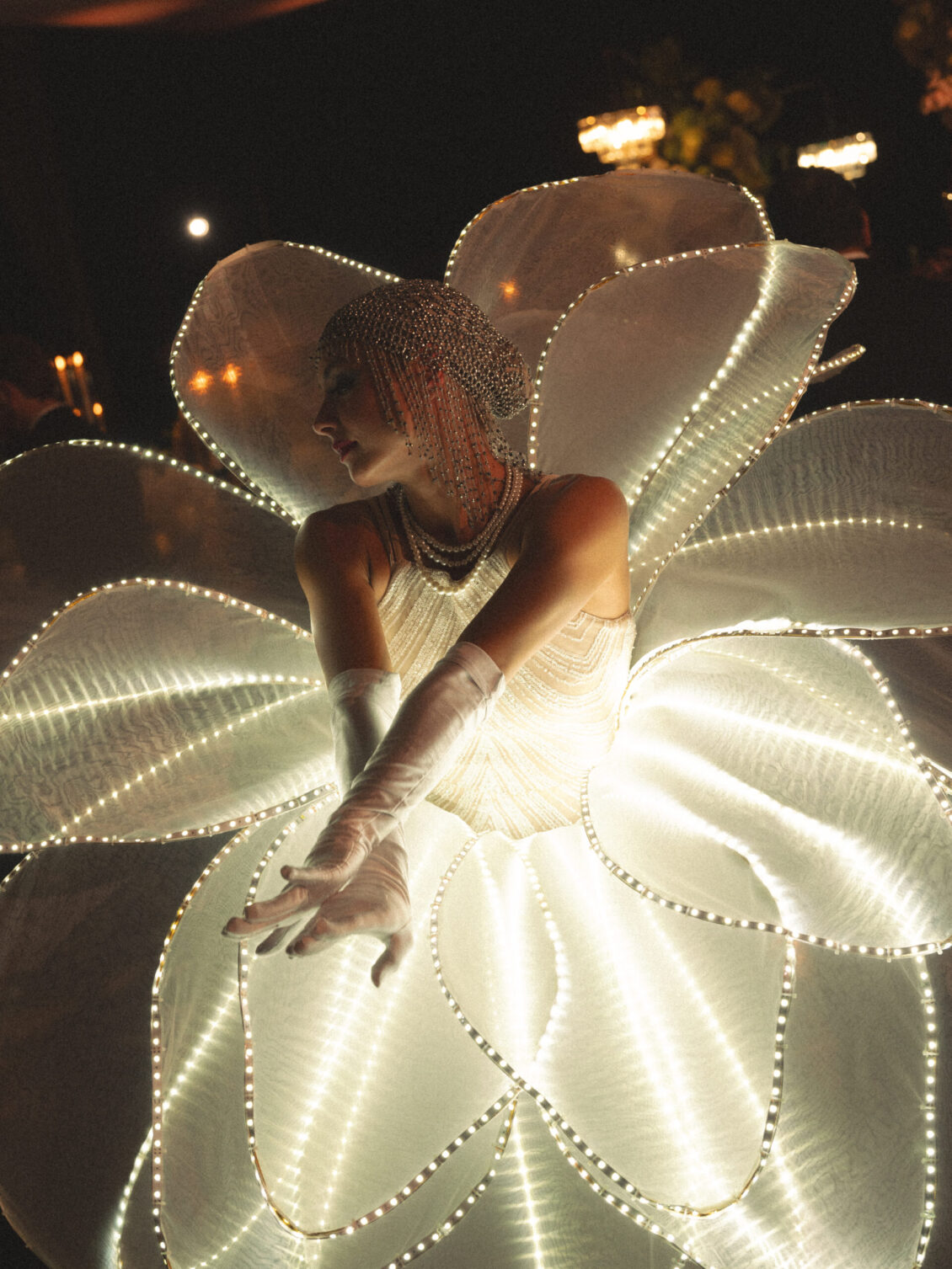 A performer bends forward with crossed arms inside a giant translucent white flower outlined by rows of small lights.
