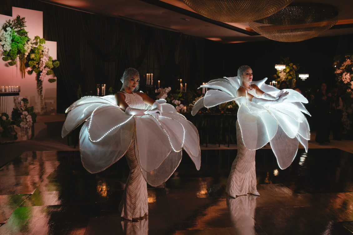 Two performers face each other inside enormous translucent white flower costumes outlined with lights on a glossy dance floor.