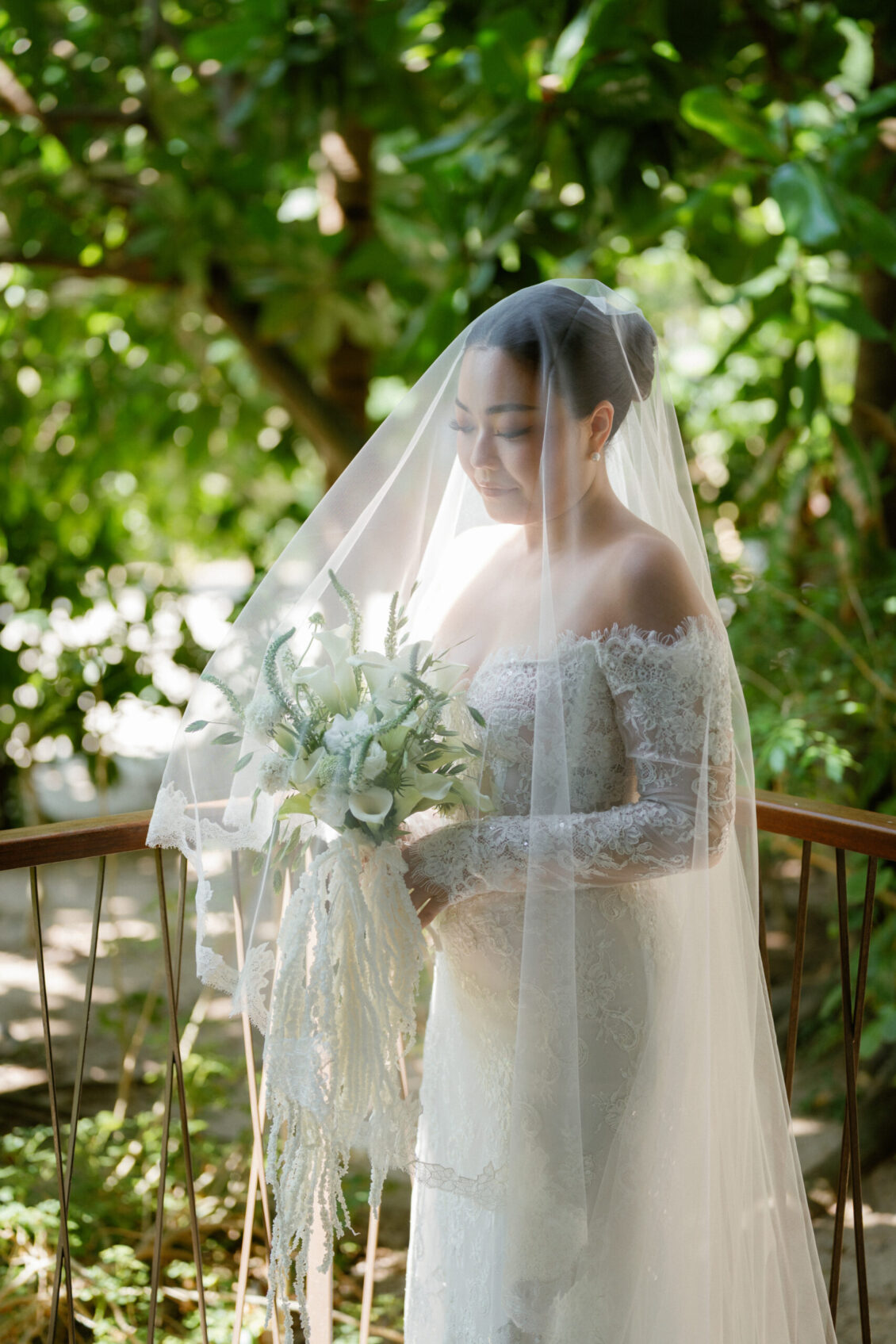 A bride in an off-shoulder lace gown stands on a wooden bridge at a Renaissance Phuket wedding, holding a bouquet of white flowers and greenery, with a sheer veil draped over her, surrounded by lush green foliage.