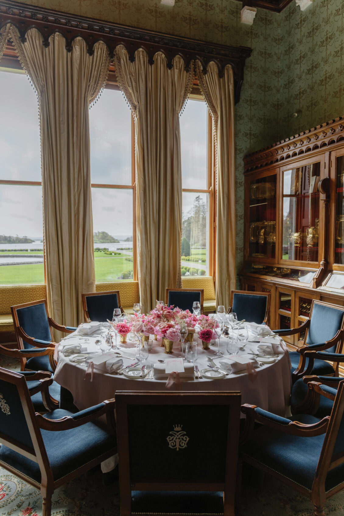 Elegant dining room with large windows, tasseled curtains, and a round table set for six with fine china and pink floral centerpieces—evoking the refined ambiance of an Irish castle wedding at Ashford Castle. Ornate wood cabinets and blue chairs complete the sophisticated setting.