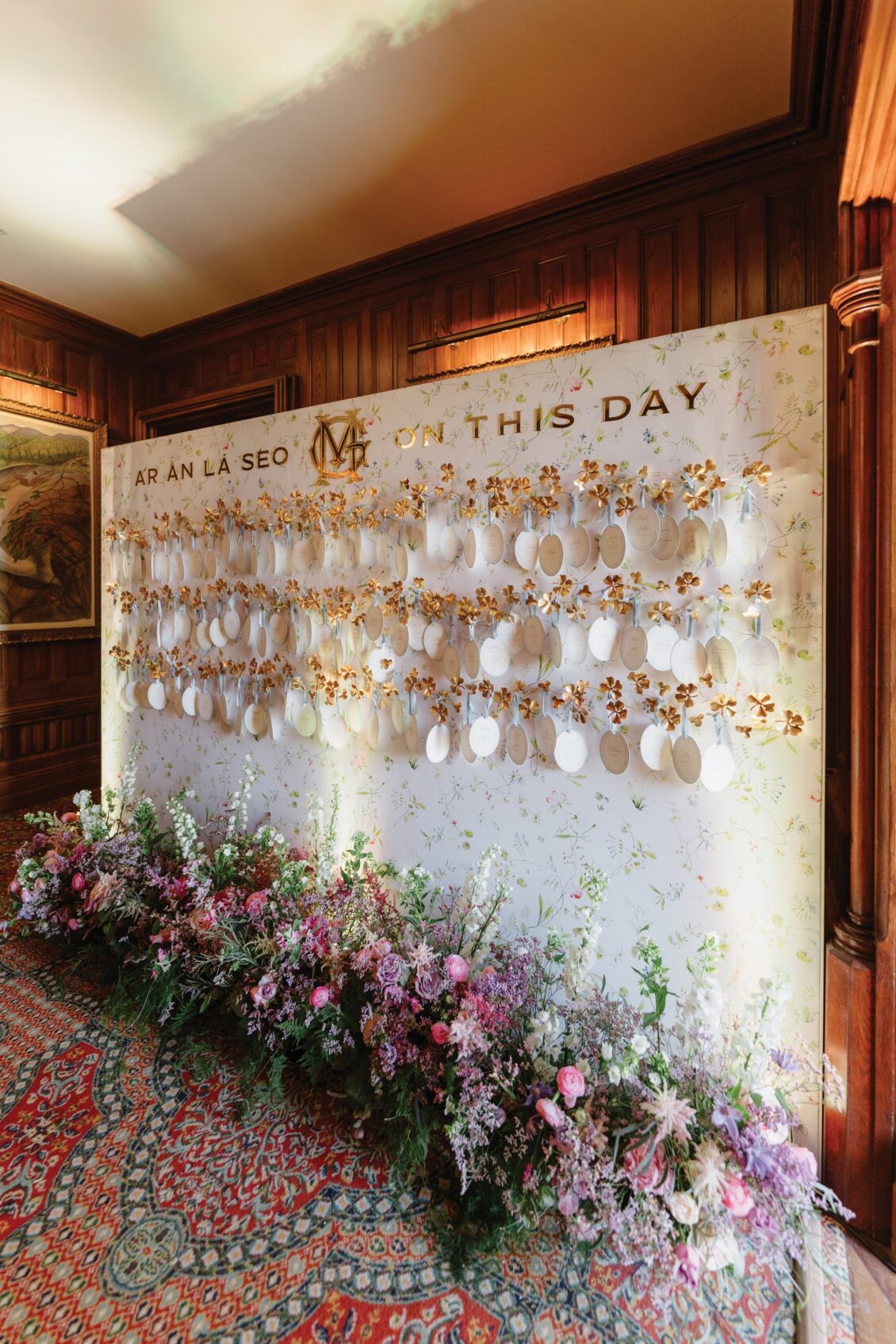 A floral-decorated board with hanging white cards and gold accents stands against a wood-paneled wall, evoking the romance of an Irish castle wedding at Ashford Castle. The board reads “AR AN LÁ SEO” and “ON THIS DAY.” Colorful flowers line the floor beneath.
