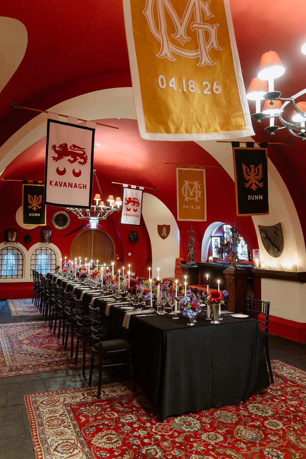 A long dining table with black chairs and candlelit settings is set in a medieval-style room with red arched ceilings, banners, shields, and stained glass windows, creating a festive, regal atmosphere reminiscent of an Irish castle wedding at Ashford Castle.