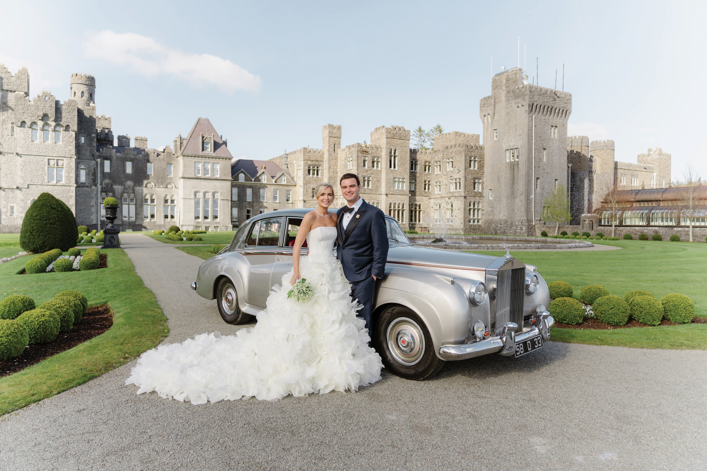 A bride and groom stand smiling beside a silver classic car in front of a large stone castle, surrounded by manicured lawns and gardens on a sunny day, capturing the elegance of an Irish castle wedding at Ashford Castle.