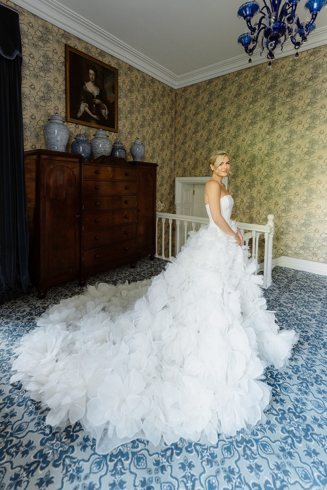 A bride in a strapless white gown with a long, ruffled train stands indoors beside a wooden dresser, under a blue chandelier, on a patterned blue carpet—capturing the timeless elegance of an Irish castle wedding at Ashford Castle.