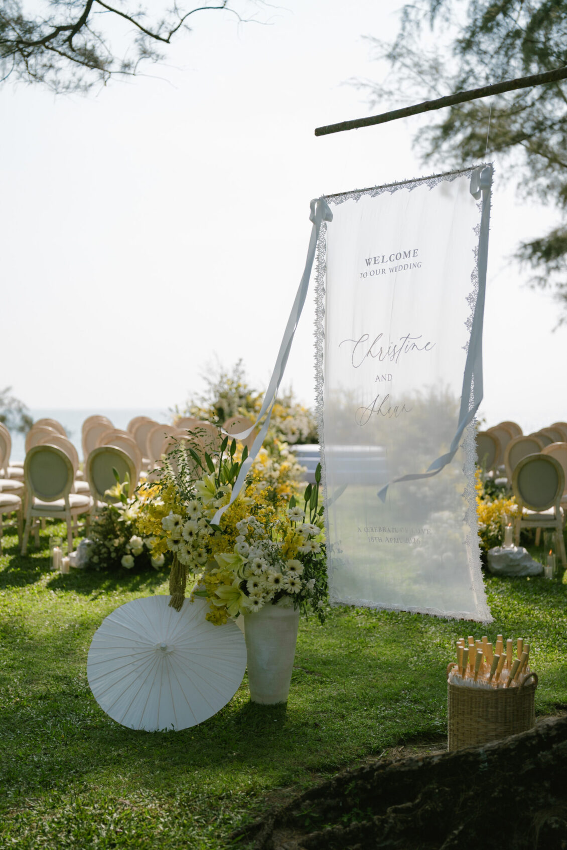 Outdoor Renaissance Phuket wedding setup with white chairs, yellow and white flowers, a transparent welcome sign, a white parasol, and a basket of fans on green grass near trees and overlooking water in the background.
