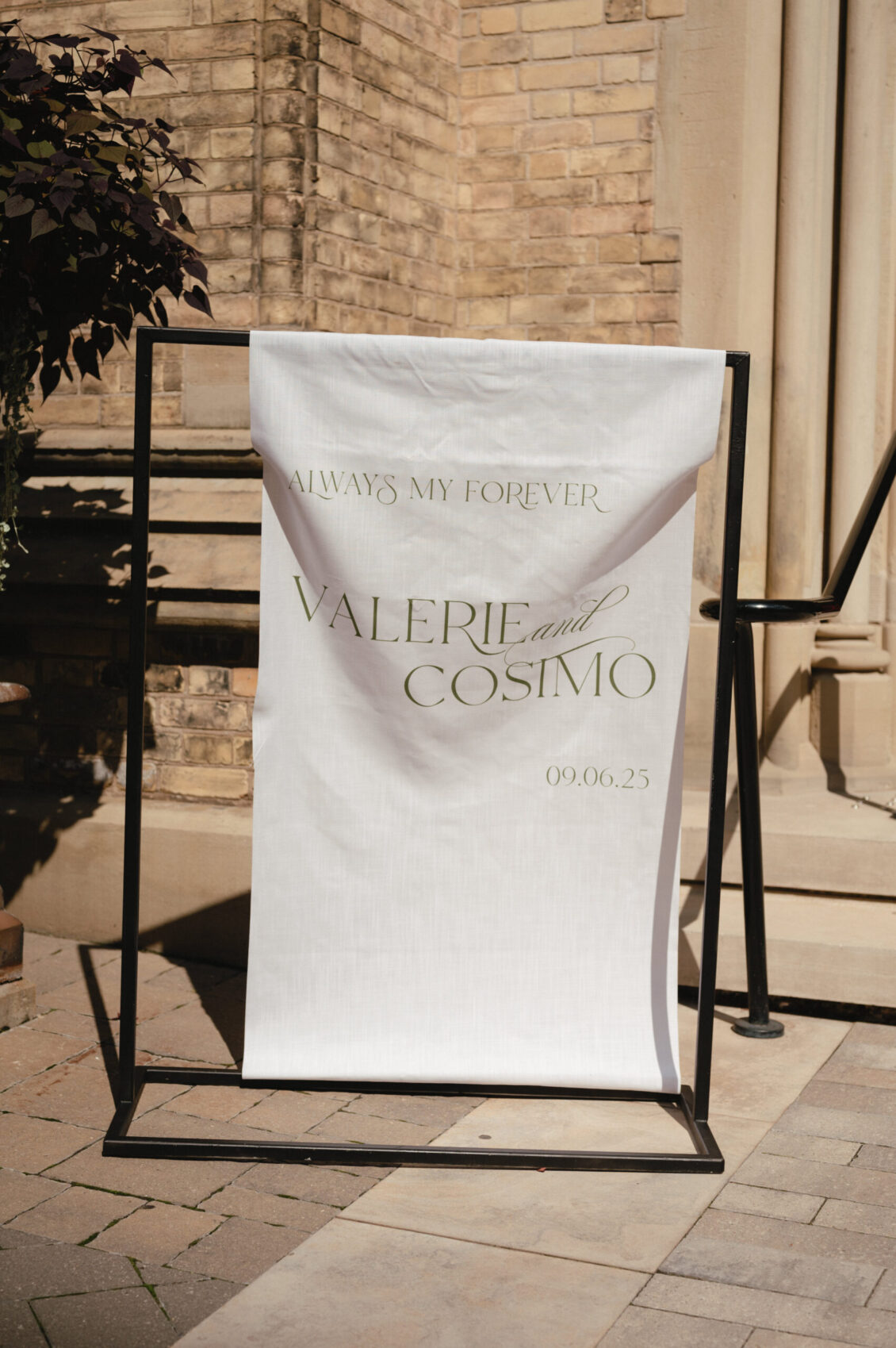 A white wedding sign reads “Always my forever. Valerie and Cosimo. 09.06.25” in elegant font, beautifully displayed on a black metal stand outside a brick building, setting the perfect scene for their black-tie wedding at Hotel X Toronto.