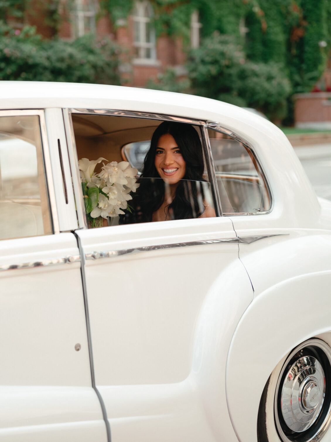 A woman in a white dress smiles while sitting in the backseat of a classic white car during a black-tie wedding at Hotel X Toronto, holding a bouquet of flowers and looking out the open window.