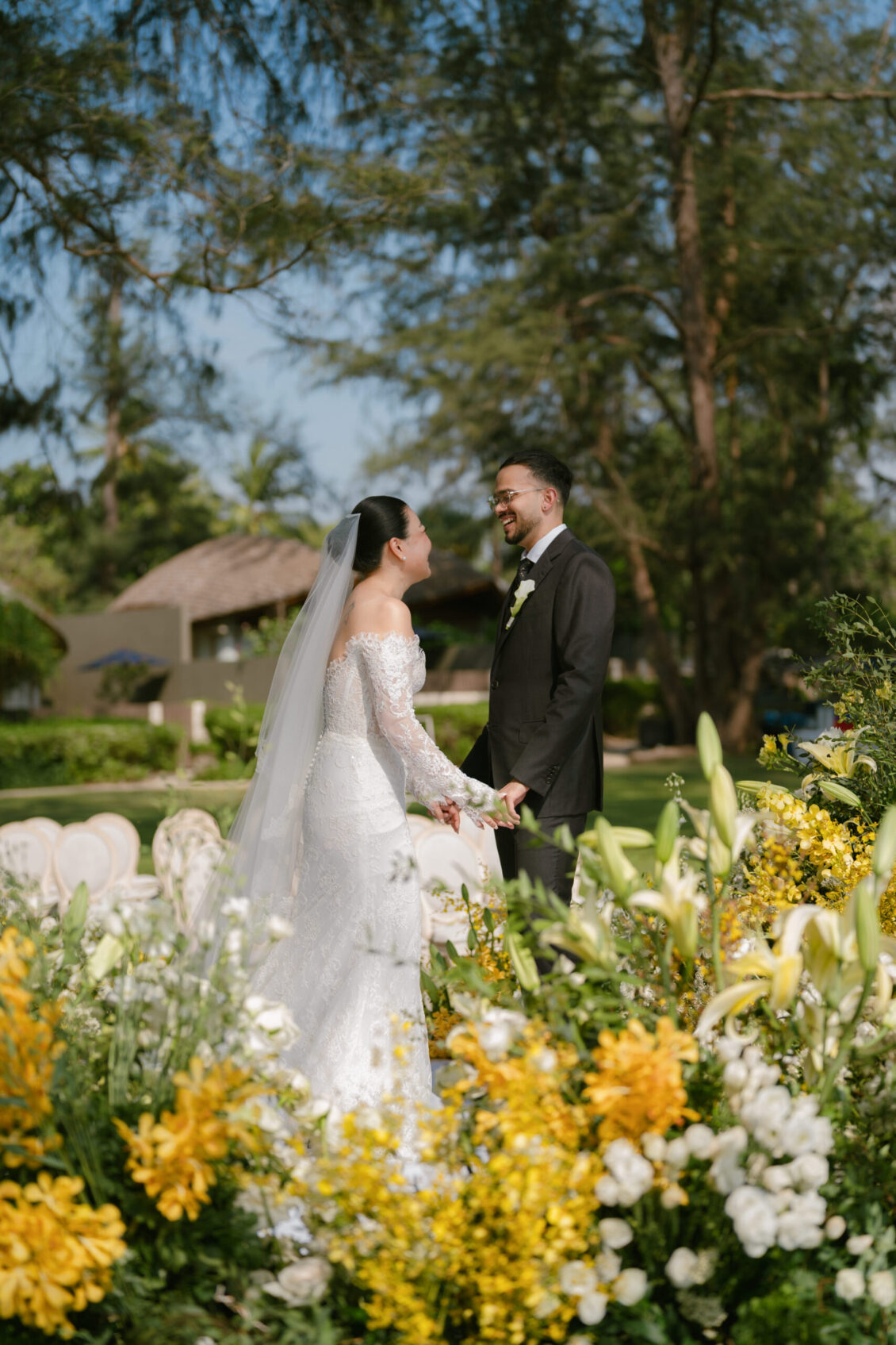 A bride and groom stand holding hands, smiling at each other, surrounded by yellow and white flowers in an outdoor garden setting at a romantic Renaissance Phuket wedding.