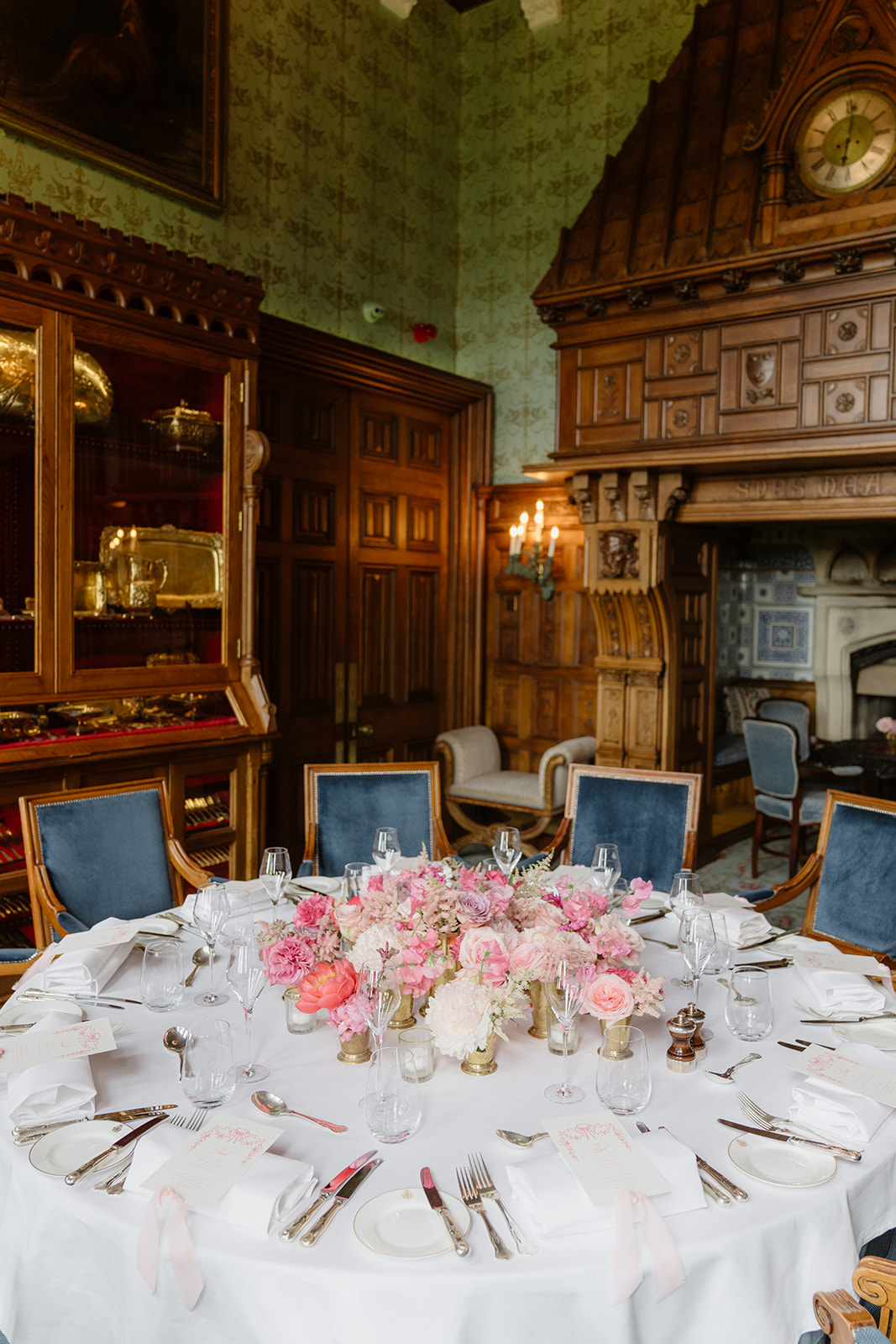 A round table set for a formal event at an Irish castle wedding at Ashford Castle features pink floral centerpieces, elegant place settings, and blue velvet chairs, arranged in a richly decorated room with wood paneling and a grand fireplace.