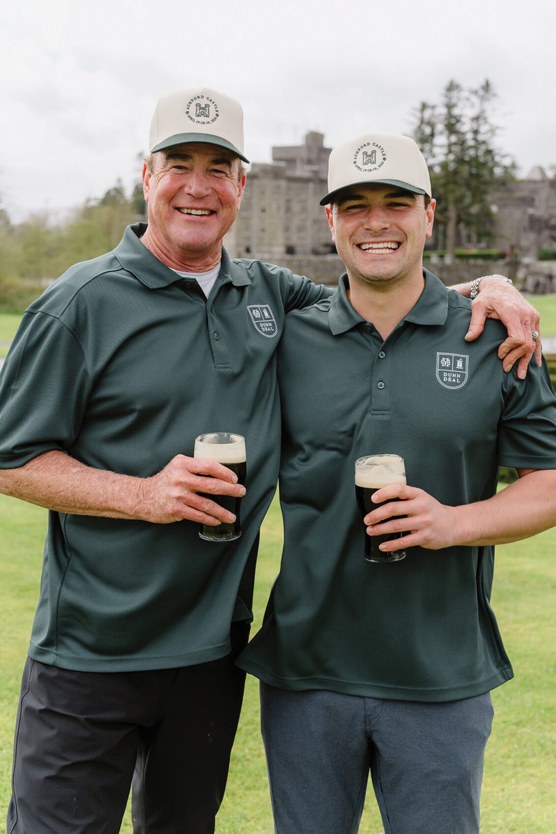 Two men in matching green shirts and hats stand on a golf course, smiling and holding pints of dark beer, with one arm around each other. Reminiscent of the camaraderie found at an Irish castle wedding at Ashford Castle, a building and trees are visible in the background.