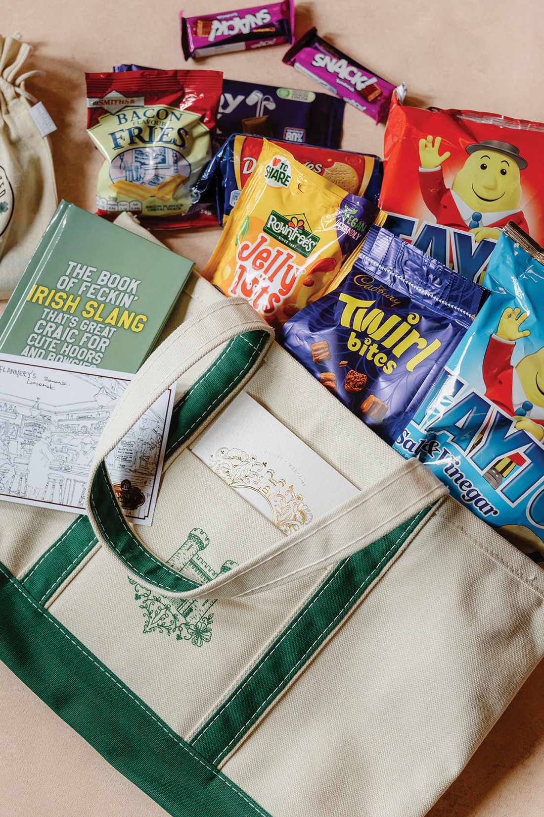 A canvas tote bag with green handles, Irish-themed designs, a book on Irish slang, greeting cards, and assorted British and Irish snacks, including crisps, chocolate, and candy, spread out on a light surface.