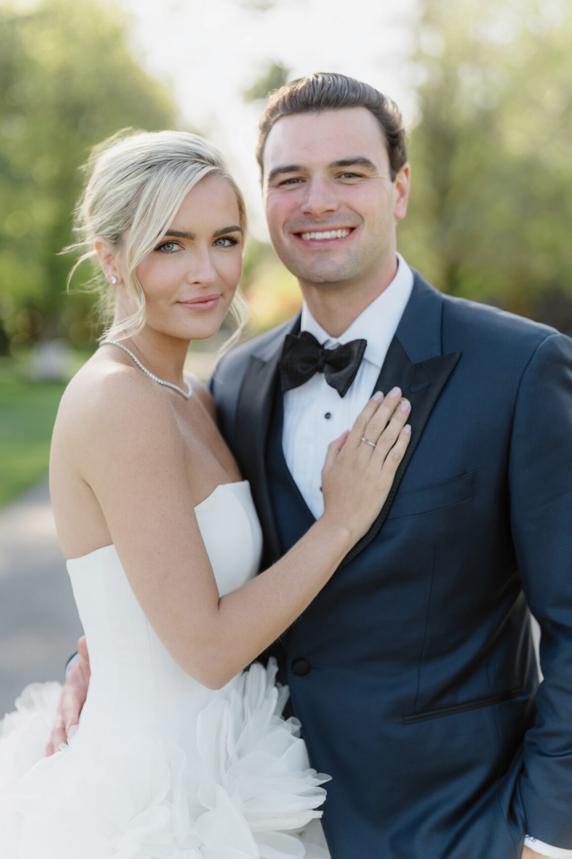 A bride in a strapless white gown and a groom in a dark tuxedo smile and pose closely together outdoors, with greenery blurred in the background, capturing the magic of their Irish castle wedding at Ashford Castle.
