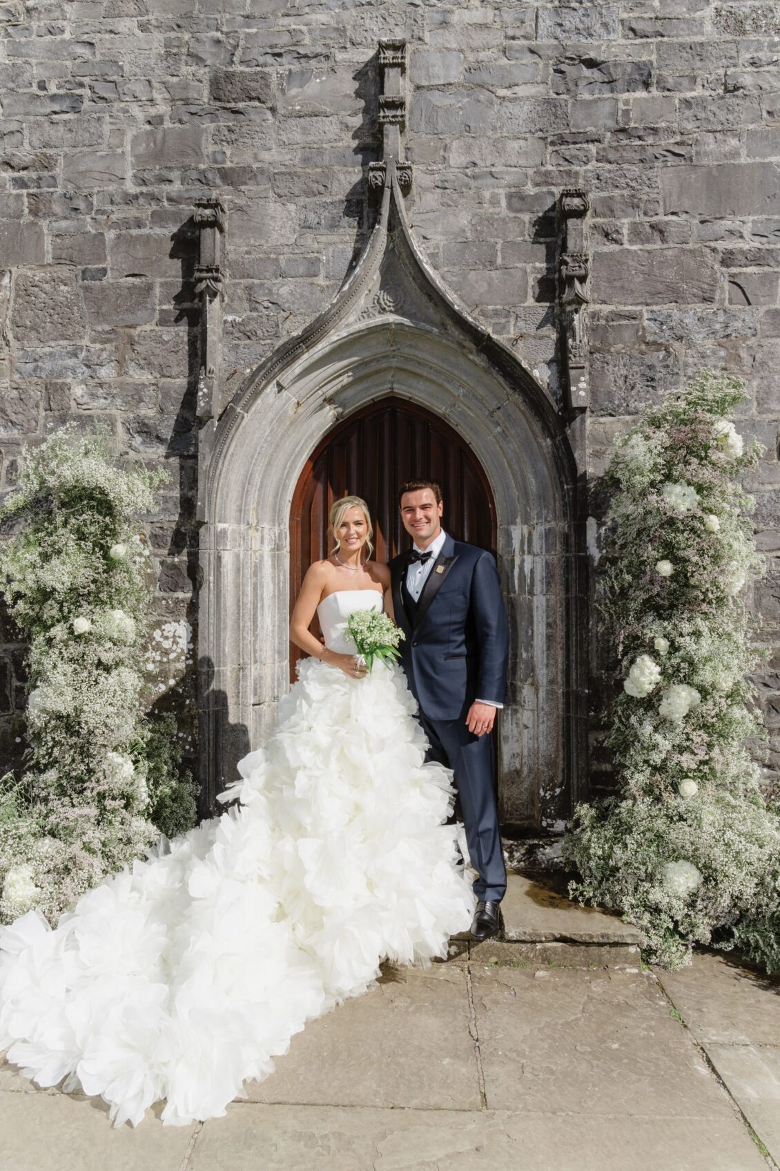A bride in a white dress and a groom in a dark suit stand smiling in front of a stone archway decorated with white flowers, capturing the romance of an Irish castle wedding at Ashford Castle.