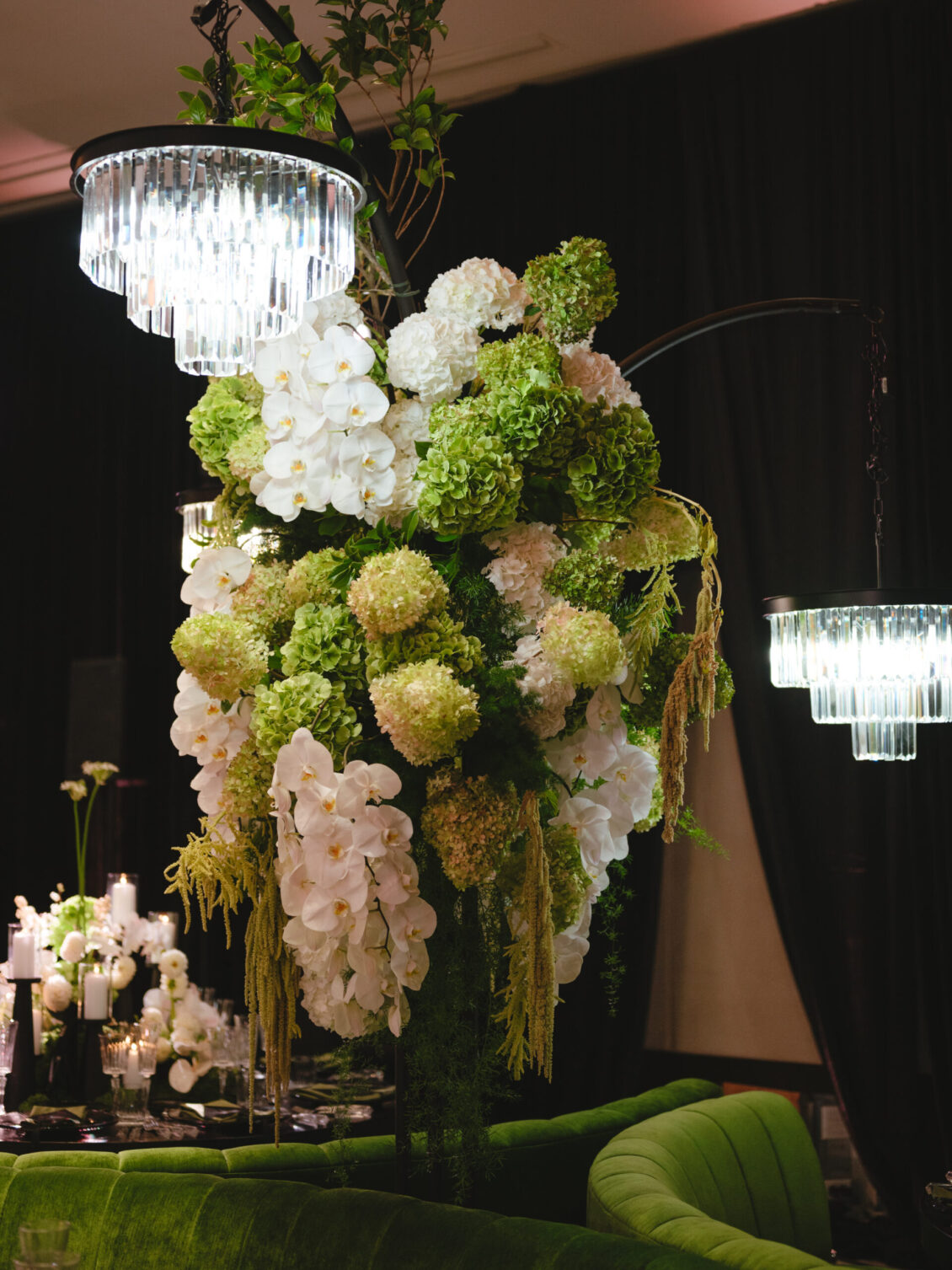 A large floral arrangement with white and green flowers hangs under crystal chandeliers, surrounded by dark curtains and green furniture—an elegant setting perfect for a black-tie wedding at Hotel X Toronto.