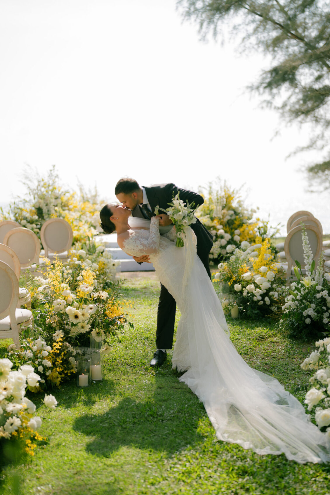 A bride and groom kiss as the groom dips the bride in a garden decorated with white and yellow flowers and empty chairs on a sunny day, capturing the romantic elegance of a Renaissance Phuket wedding.