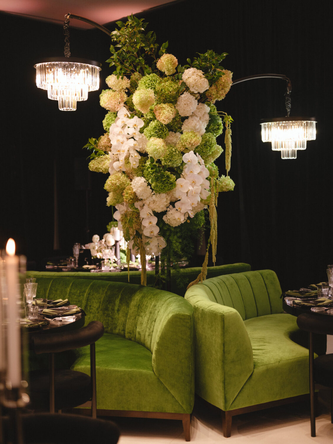 Lush green velvet sofas surround a table, topped with a large floral arrangement of white and green blooms. Two elegant chandeliers hang above, creating a luxurious, intimate dining space with candlelight—perfect for a black-tie wedding at Hotel X Toronto.