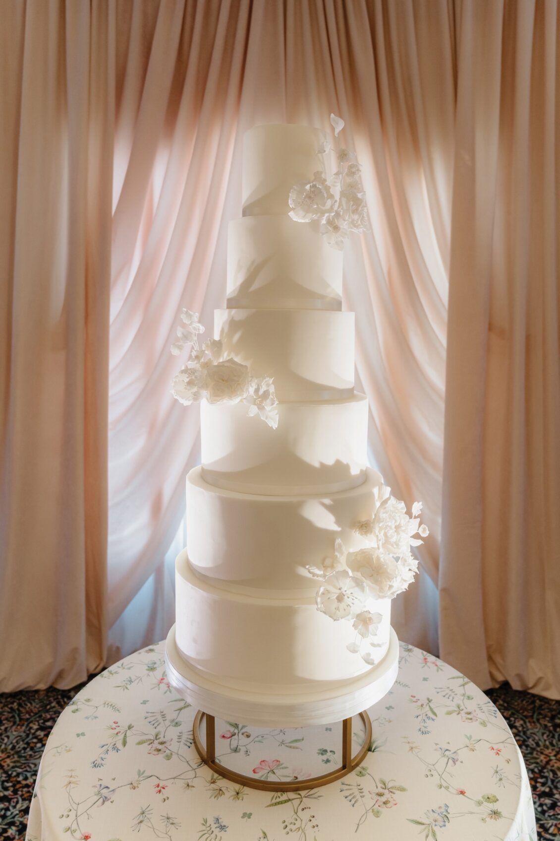 A tall, white, six-tier wedding cake with delicate white floral decorations stands on a round table, set against soft, draped pink curtains—an elegant centerpiece perfectly suited for an Irish castle wedding at Ashford Castle.