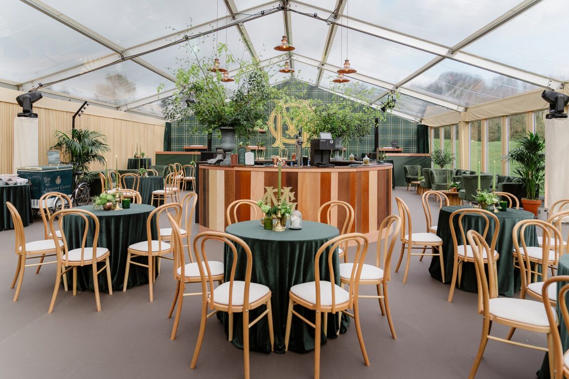 Event tent with round tables covered in green cloth, wooden chairs, and a central bar. Greenery and plants decorate the space, and large windows let in natural light. Elegant, festive, and ready for guests—perfect for an Irish castle wedding at Ashford Castle.