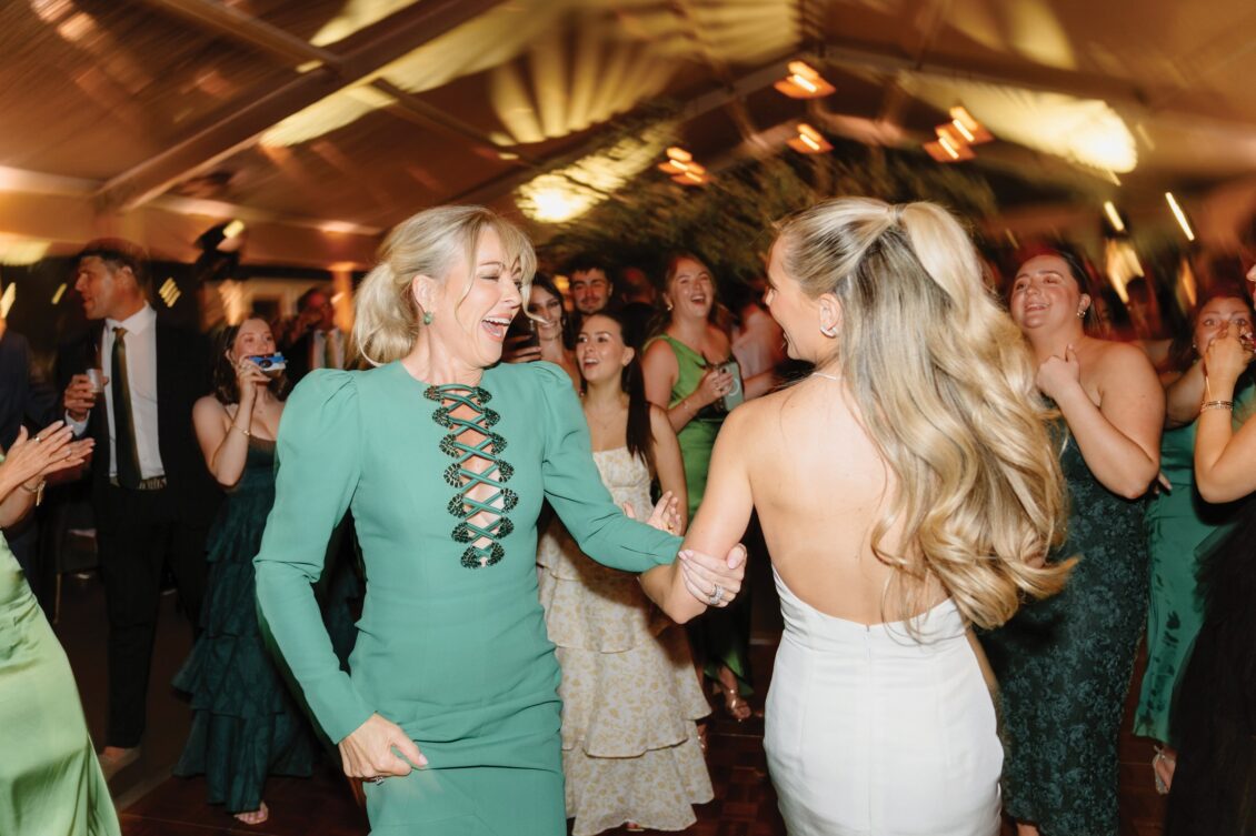Two women, one in a green dress and one in a white strapless dress, dance joyfully at a lively indoor celebration surrounded by cheering guests in green attire during an Irish castle wedding at Ashford Castle.