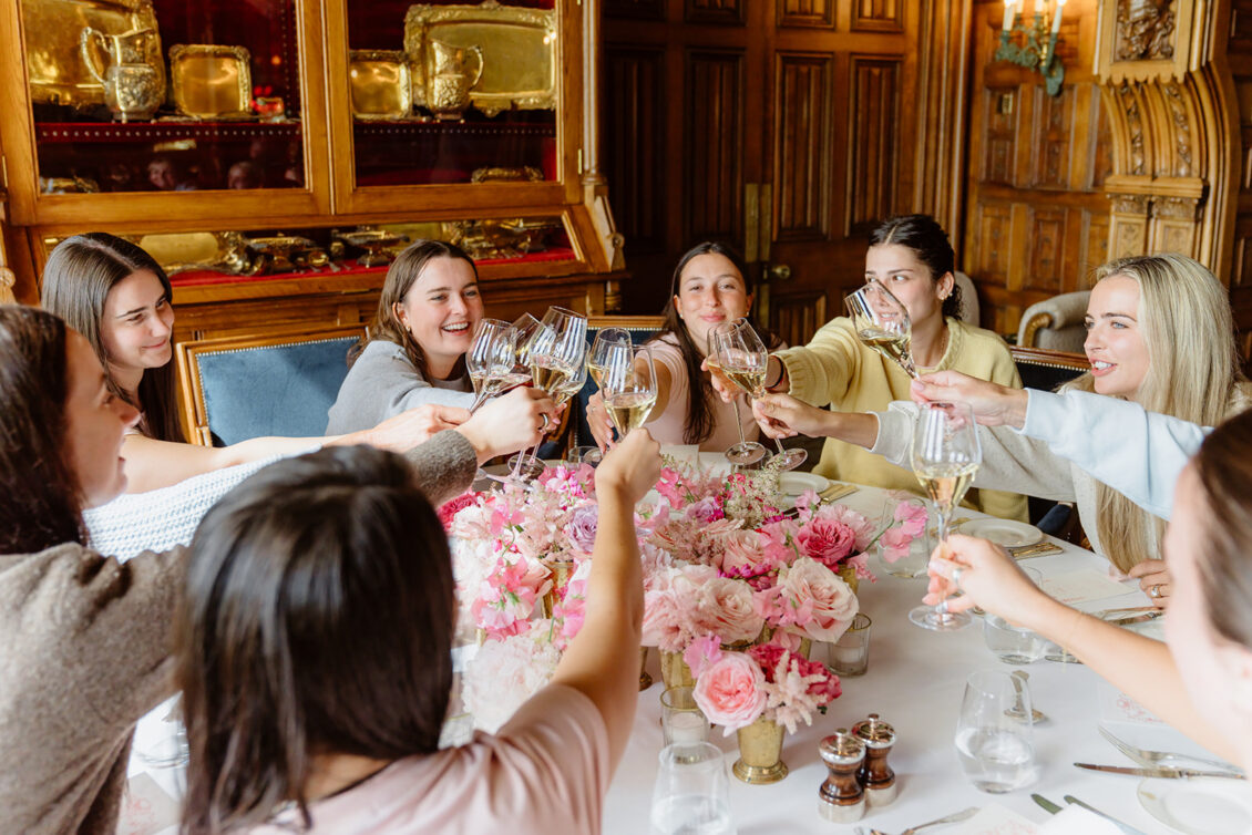 Seven women sit around a table with pink flowers, raising glasses in a toast and smiling in a wood-paneled, ornate room at an Irish castle wedding at Ashford Castle.
