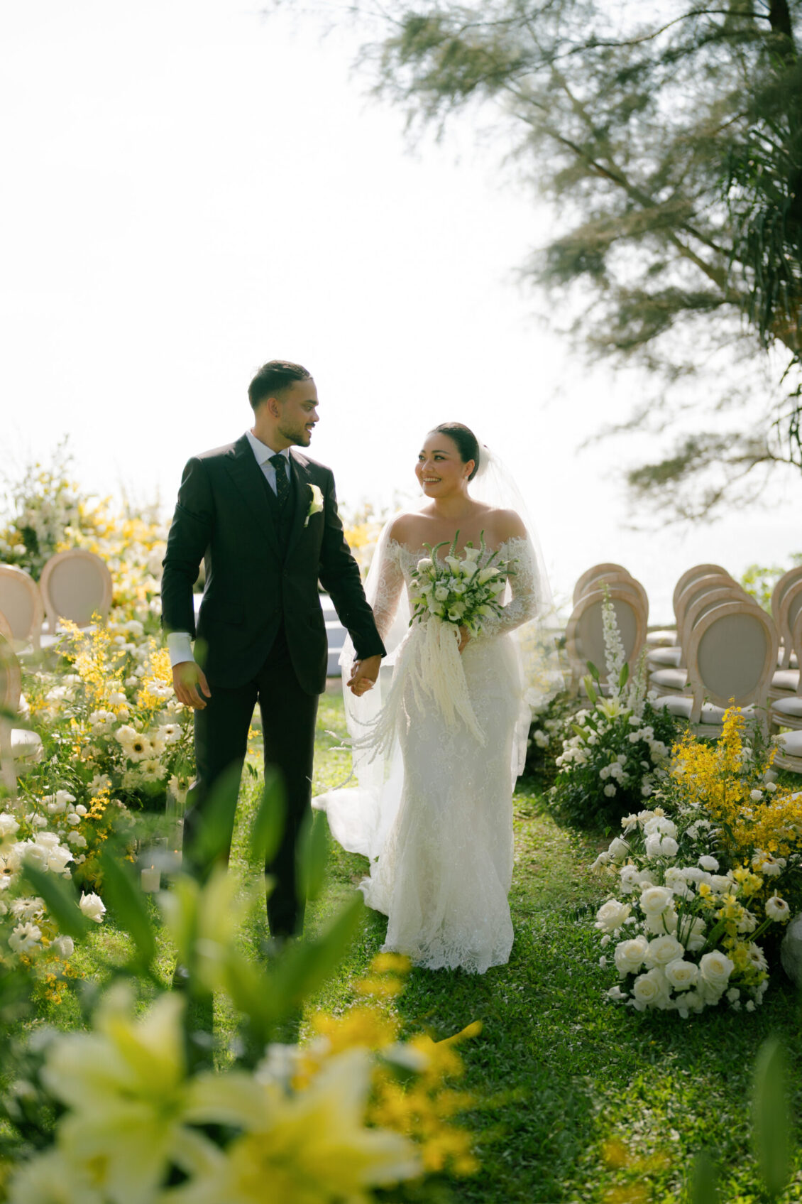 A bride and groom walk hand in hand down an outdoor aisle lined with yellow and white flowers, smiling at each other on their Renaissance Phuket wedding day.