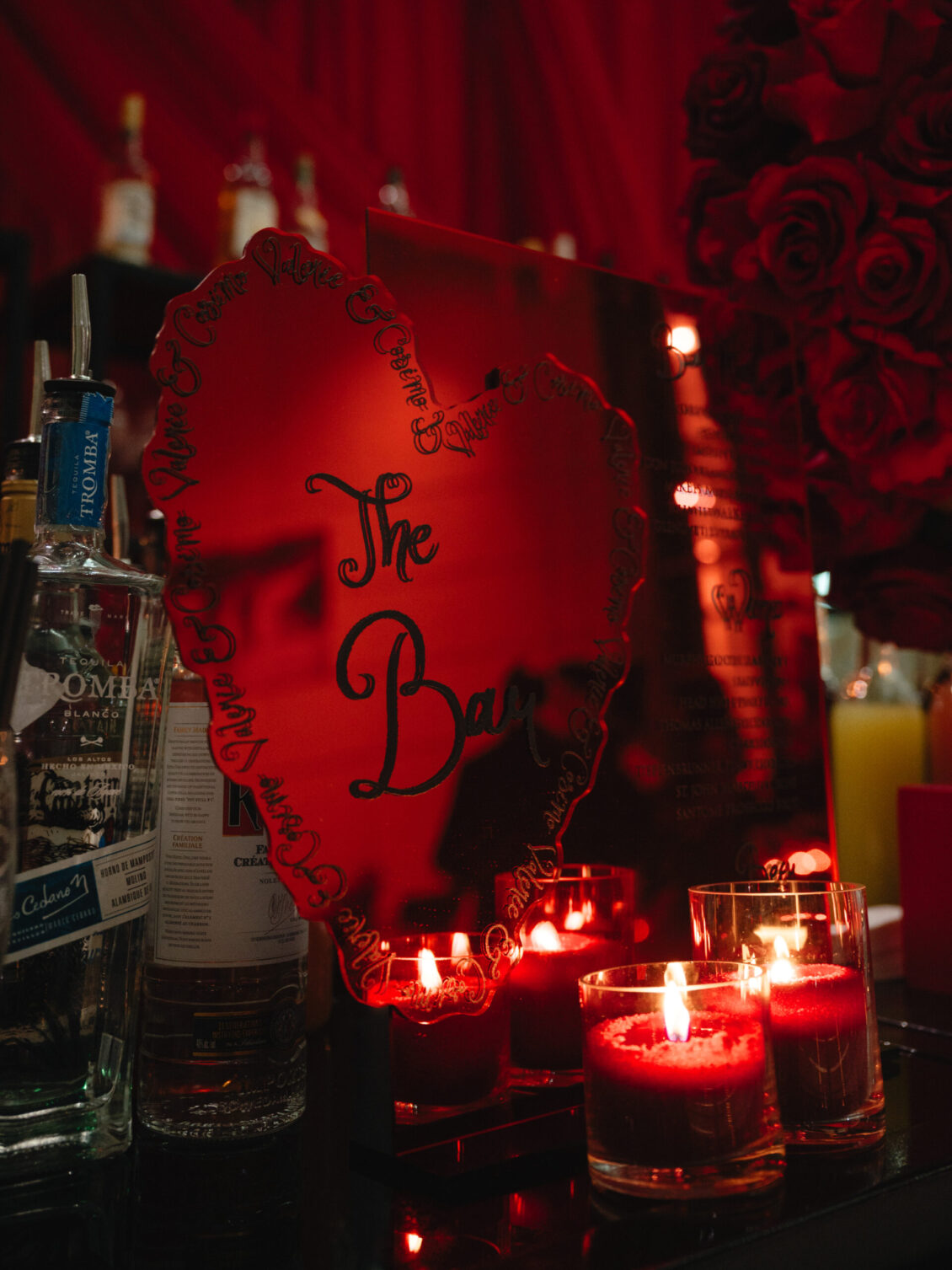A heart-shaped bar sign with “The Bar” written on it, surrounded by various liquor bottles, red roses, and lit red candles, creating a romantic and elegant atmosphere reminiscent of a black-tie wedding at Hotel X Toronto.