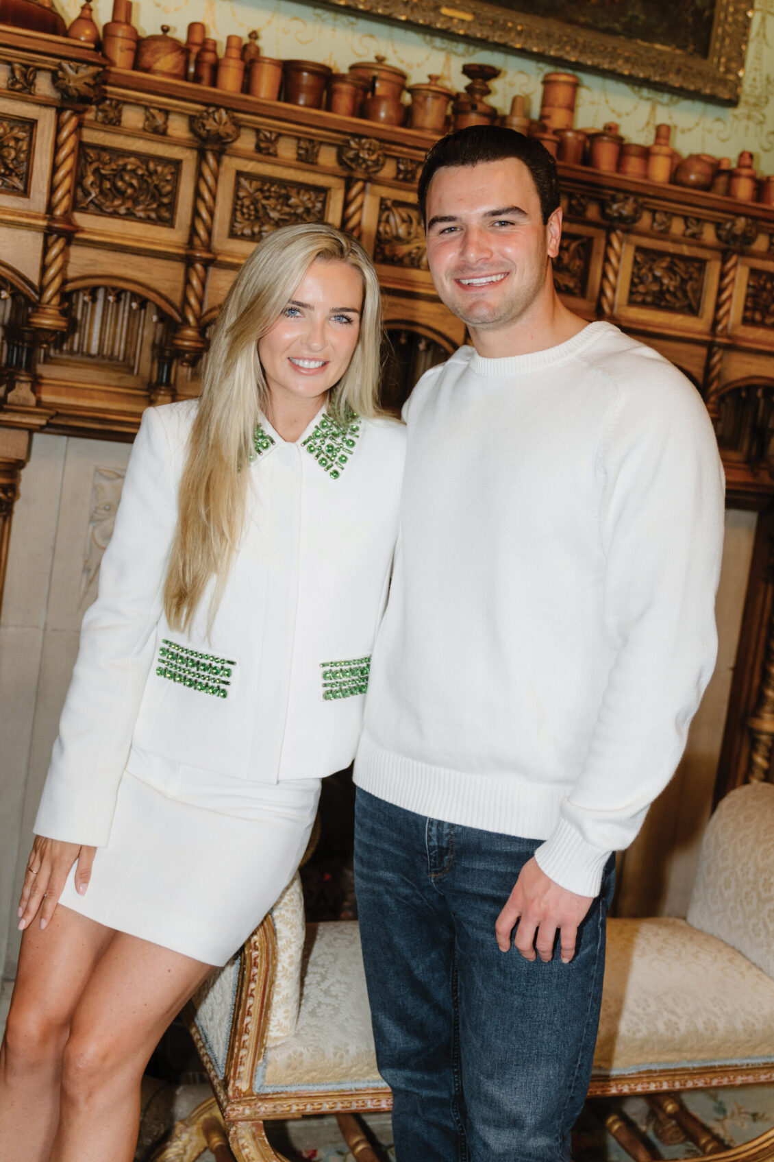 A woman in a white outfit with green details stands beside a man in a white sweater and jeans. They both smile, posing in an ornate, elegant room with wooden paneling and decorative details, evoking the timeless charm of an Irish castle wedding at Ashford Castle.