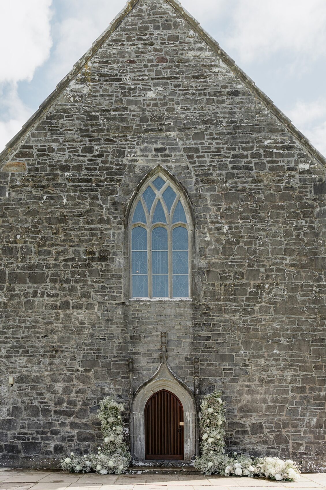 A stone church facade with a pointed arched window above a small wooden door, framed by white flowers at the entrance, evokes the timeless romance of an Irish castle wedding at Ashford Castle. The sky and some clouds are visible above the roof, adding to the enchanting atmosphere.