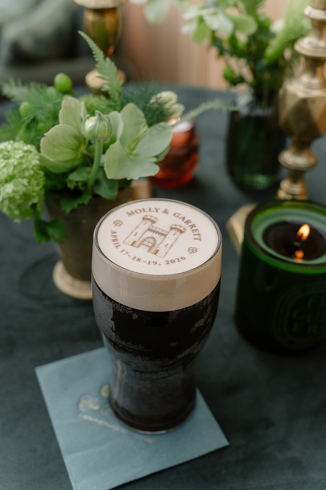 A pint of dark beer with custom foam art reading “Molly & Garrett” and a date, on a napkin, surrounded by green plants, a candle, and brass decor—capturing the timeless romance of an Irish castle wedding at Ashford Castle.