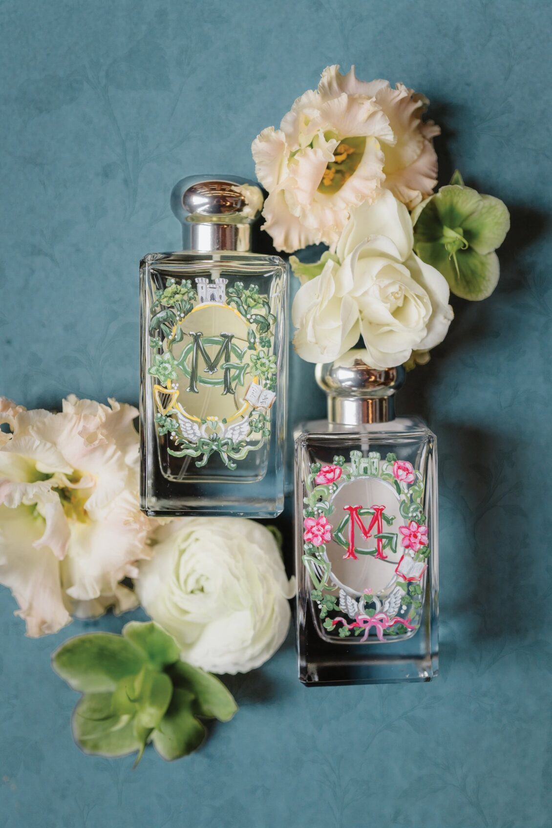 Two elegant perfume bottles with ornate, floral labels are placed on a blue surface, surrounded by soft white and pale pink flowers and green leaves—evoking the romance of an Irish castle wedding at Ashford Castle.