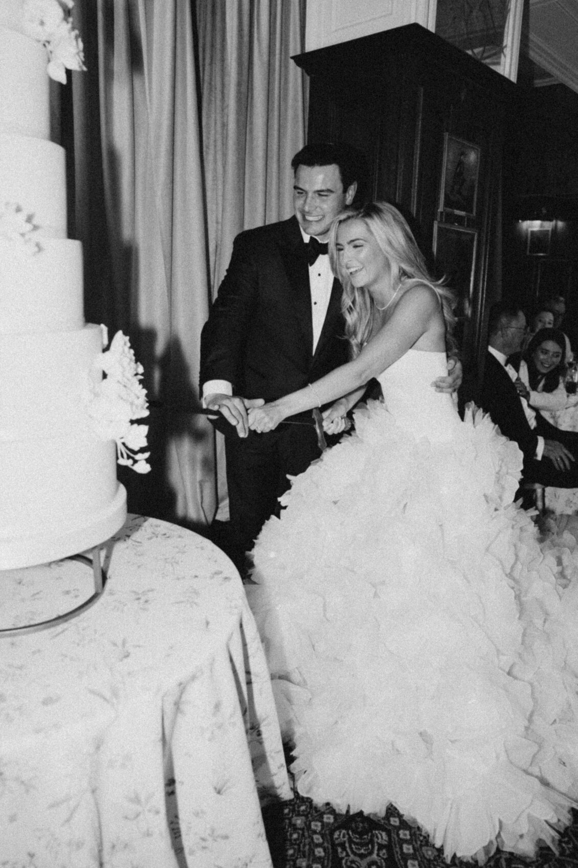 A bride and groom, smiling in formal wedding attire, cut a tiered white wedding cake together in the grand hall of their Irish castle wedding at Ashford Castle, with guests seated in the elegantly decorated indoor setting.