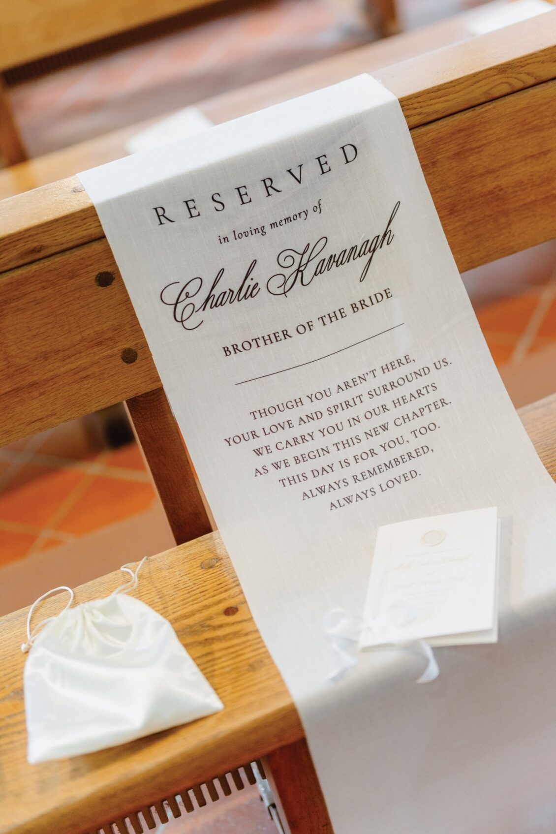 A white memorial sign on a church pew at this Irish castle wedding at Ashford Castle reads, “Reserved in loving memory of Charlie Kavanagh, brother of the bride,” with a heartfelt message below. A small white bag and card are placed on the pew.