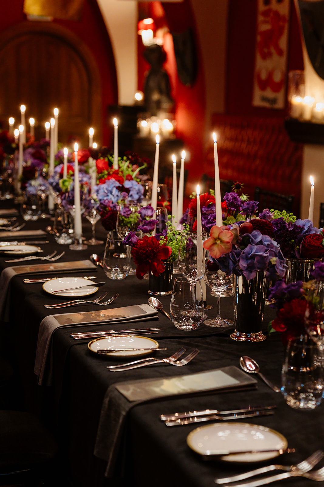 A long banquet table with a black tablecloth, set with plates, cutlery, wine glasses, tall white candles, and colorful flower arrangements in a dim, elegant room—perfectly capturing the enchanting atmosphere of an Irish castle wedding at Ashford Castle.