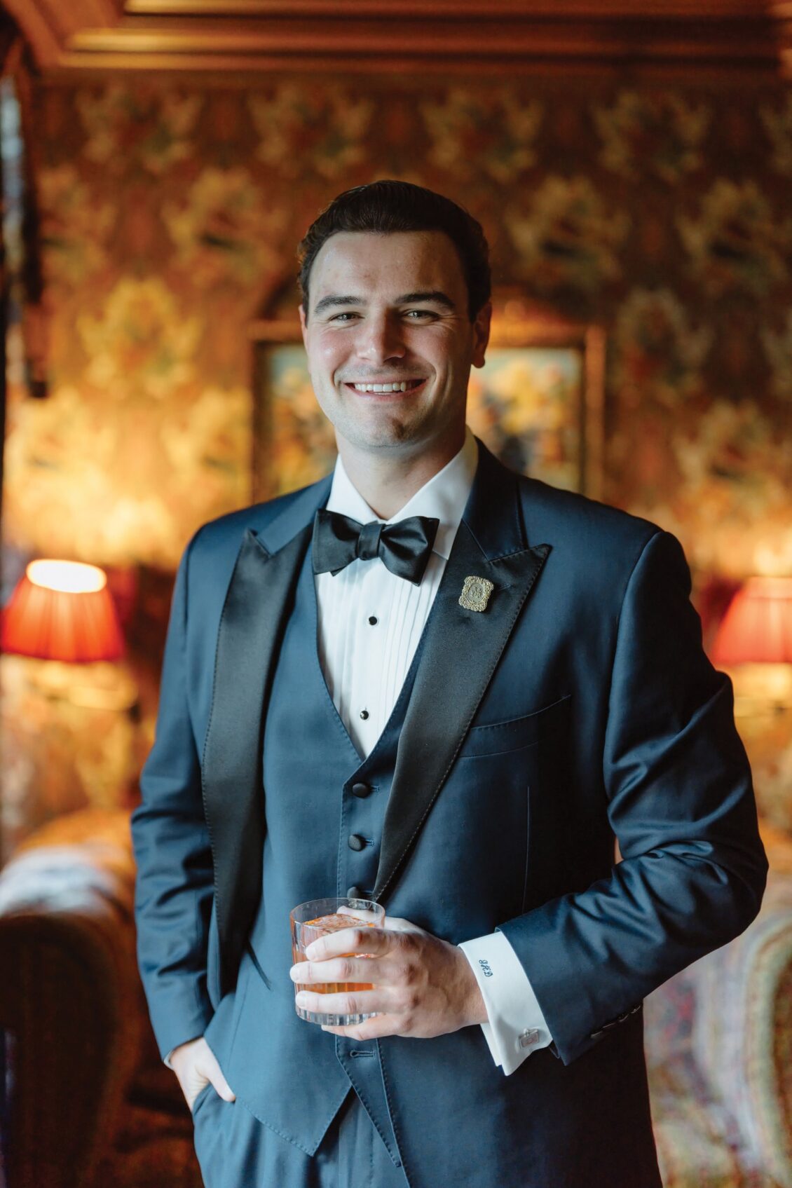A smiling man in a navy tuxedo with a bow tie stands indoors, holding a drink in one hand. The background includes patterned wallpaper, red lamps, and soft lighting, evoking the elegant ambiance of an Irish castle wedding at Ashford Castle.