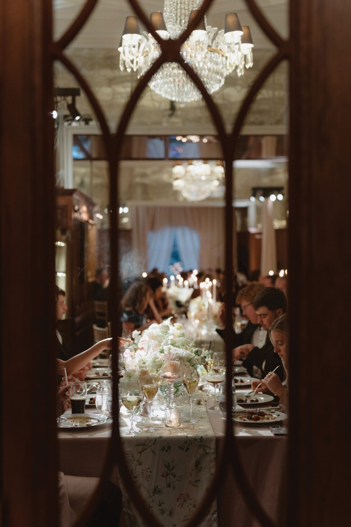 Elegant dinner party seen through a decorative glass door, reminiscent of an Irish castle wedding at Ashford Castle, with guests seated at a long, flower-adorned table under chandeliers, engaged in conversation and dining.