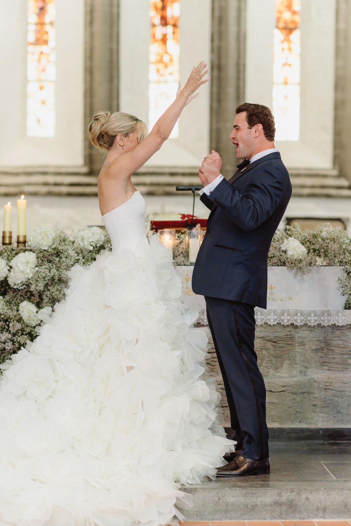 A bride in a ruffled white dress raises her arms excitedly while facing a groom in a dark suit. They appear joyful, standing at an altar decorated with flowers and candles inside the bright halls of an Irish castle wedding at Ashford Castle.