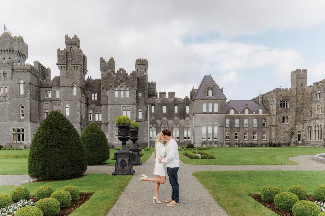 A couple embraces and kisses in front of a large stone castle, surrounded by manicured gardens and cloudy skies, capturing the romance of an Irish castle wedding at Ashford Castle.