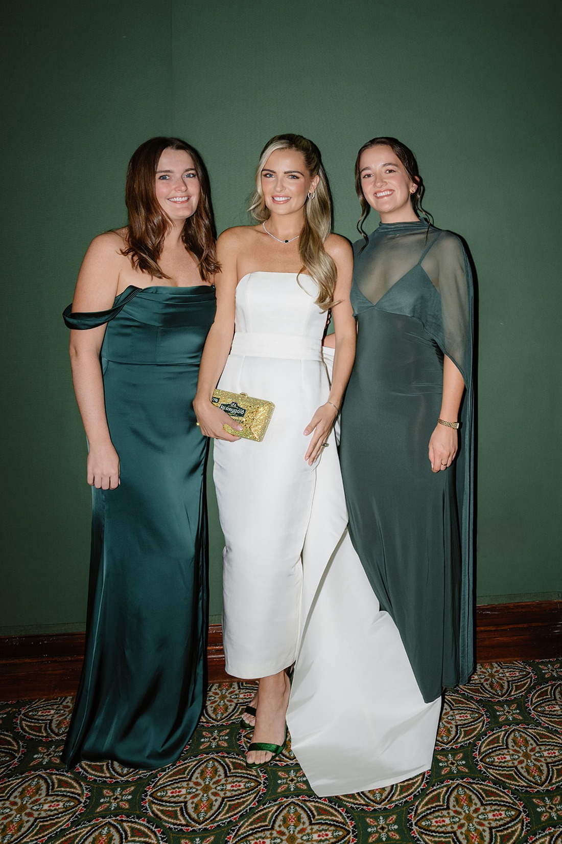 Three women in formal evening dresses pose and smile in front of a green wall at an Irish castle wedding at Ashford Castle. Two wear dark green gowns; the one in the middle wears a white strapless dress and holds a sparkly gold clutch.