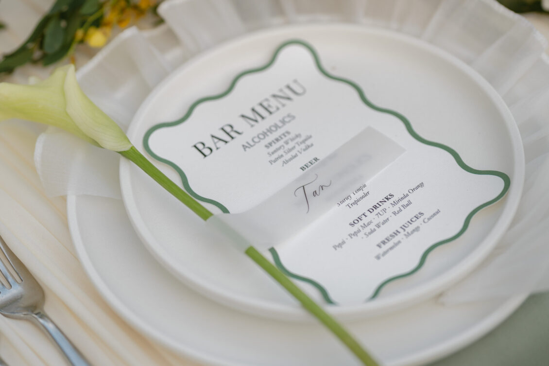 A close-up of a white plate with a bar menu card placed on top at a Renaissance Phuket wedding. A yellow calla lily flower rests on the plate, and a fork is seen to the left. The menu lists alcoholic and non-alcoholic drink options.