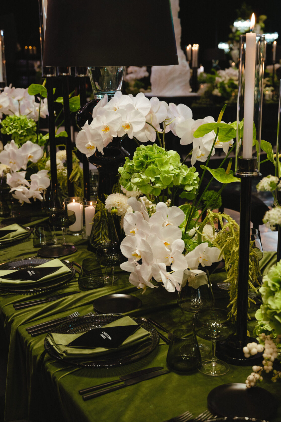 An elegant dining table, perfect for a black-tie wedding at Hotel X Toronto, features a green tablecloth, black tableware, tall black candlesticks, and lush floral arrangements of white orchids and green hydrangeas.