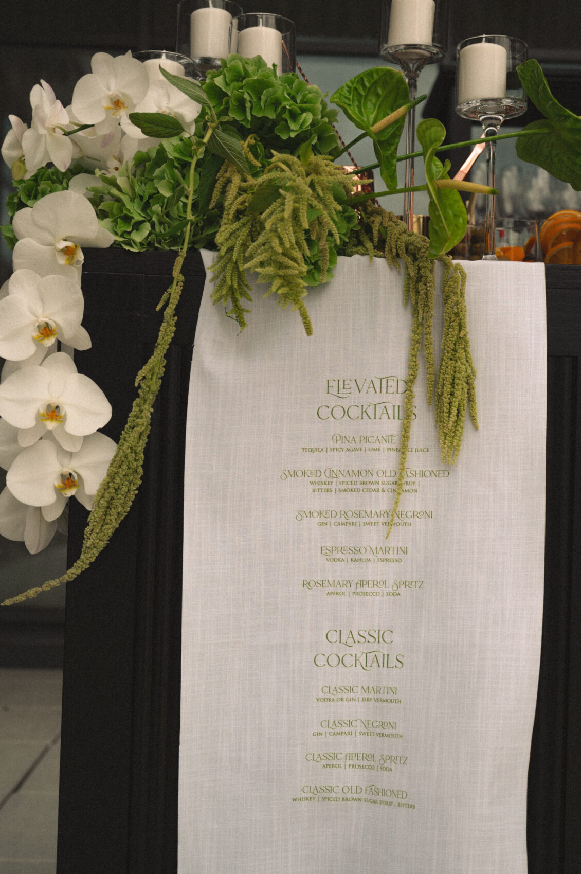 A linen cocktail menu with a floral and greenery arrangement above it, listing elevated and classic cocktails in gold text, sets an elegant scene at this black-tie wedding at Hotel X Toronto. Candles in glass holders are seen on a dark table behind the menu.
