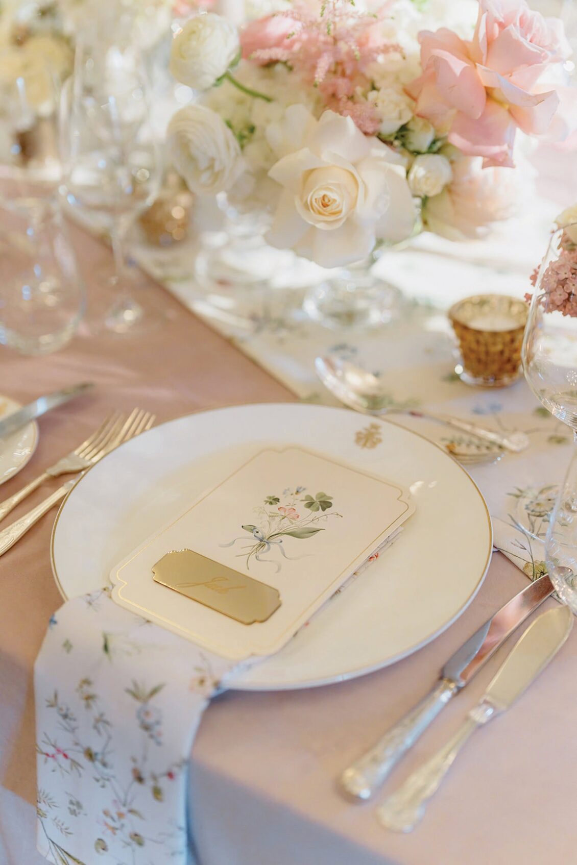 Elegant table setting with a white plate, floral napkin, menu card, and gold place card. Surrounding the plate are silver cutlery, crystal glasses, and a pastel floral centerpiece. Soft, romantic ambiance sets the tone for an unforgettable Irish castle wedding at Ashford Castle.