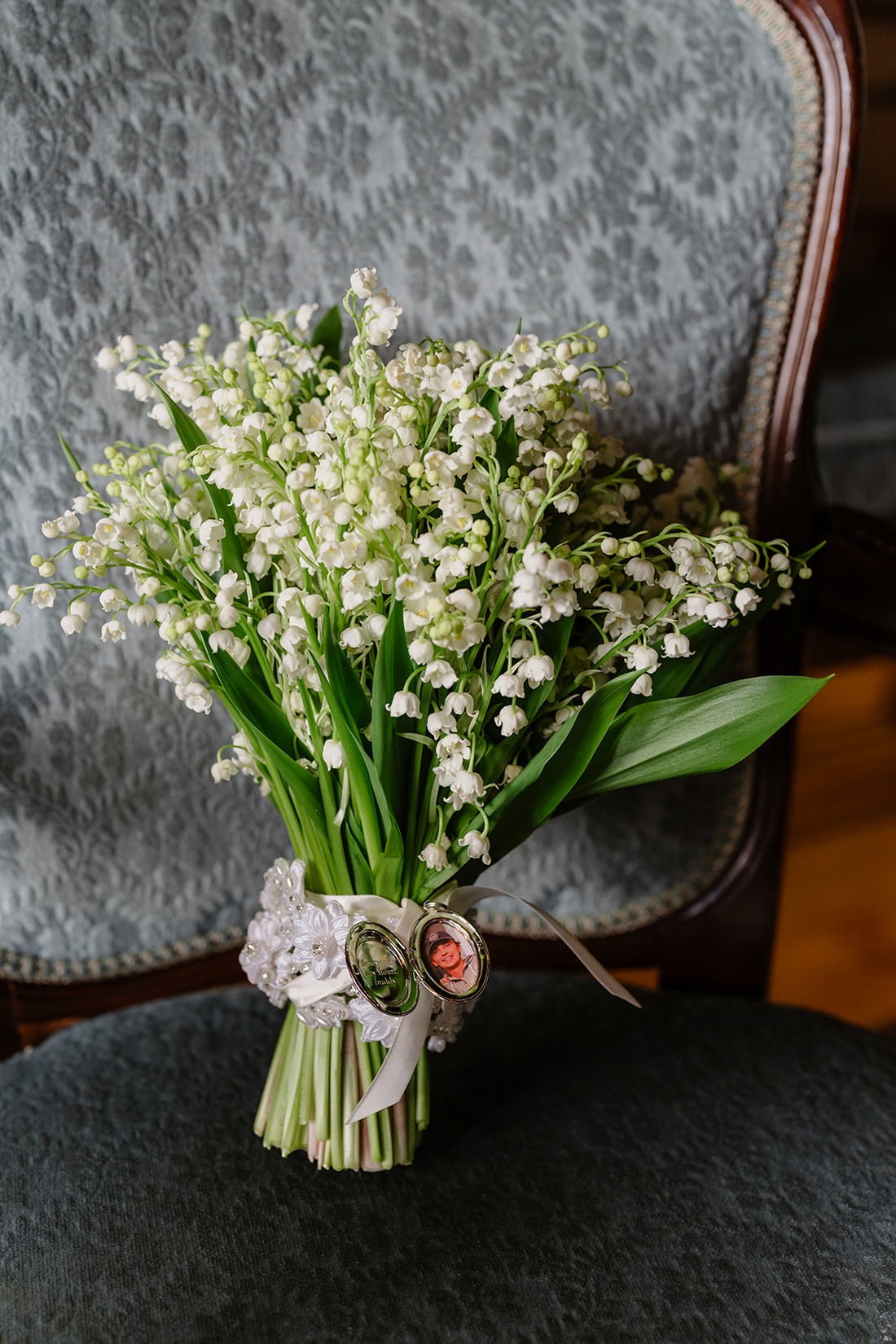 A bouquet of lily of the valley flowers with green leaves, tied with a white ribbon and adorned with vintage-style photo charms, rests on a gray patterned chair—an elegant detail befitting an Irish castle wedding at Ashford Castle.