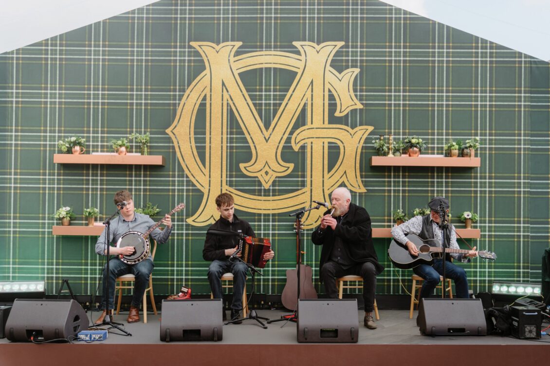 Four musicians sit on stage in front of a large gold monogram on a green plaid backdrop, playing banjo, accordion, flute, and guitar—with potted plants on shelves behind them—setting the enchanting atmosphere for an Irish castle wedding at Ashford Castle.