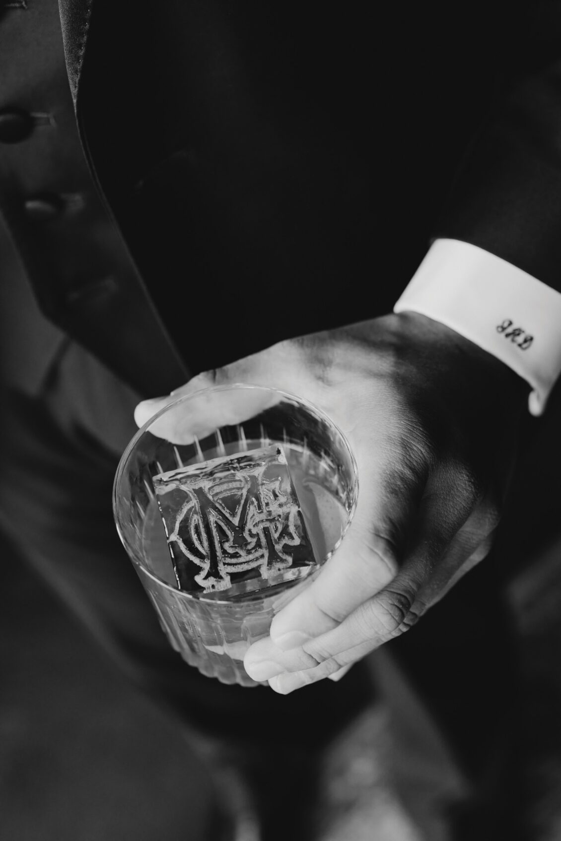 A person in a suit holds a glass with a large square ice cube engraved with initials, perhaps toasting at an Irish castle wedding at Ashford Castle. Their cufflink shows the number “342.” The image is black and white.