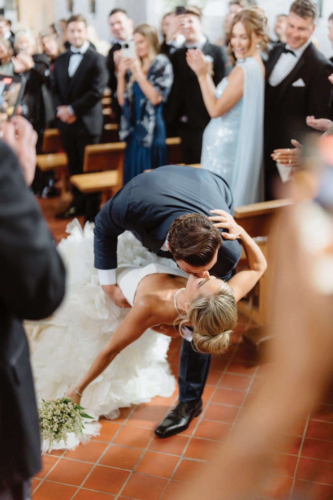 A groom dips his bride for a kiss in the aisle of an Irish castle wedding at Ashford Castle, surrounded by applauding and smiling wedding guests dressed in formal attire.