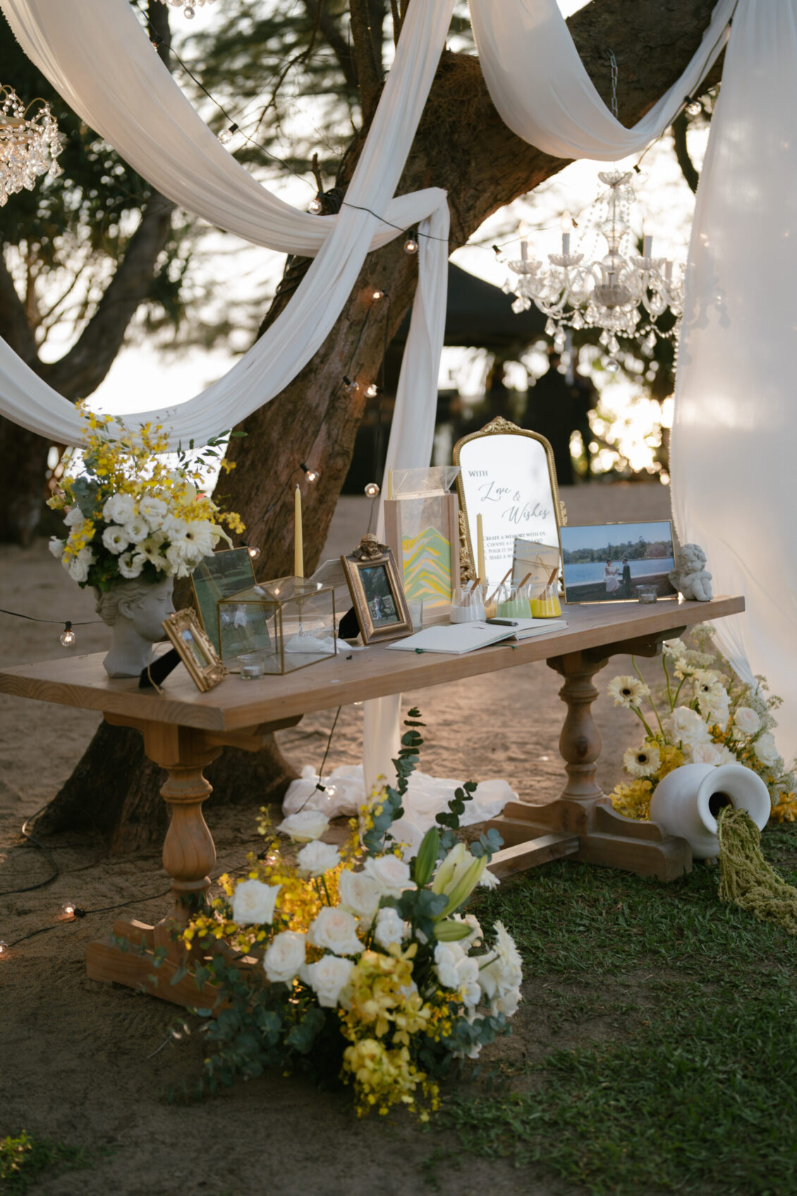 A decorated wooden table outdoors with framed photos, candles, and yellow and white flowers, under draped white fabric and a crystal chandelier, creating a romantic, elegant setting reminiscent of a Renaissance Phuket wedding.
